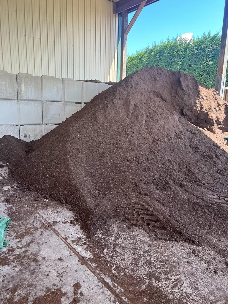 A large pile of rich, dark soil stored under a shelter at Newcomb's Nursery, essential for the cultivation and potting of various plant species.
